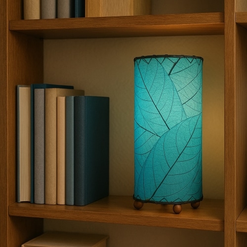 Eangee Home Design Mini Cylinder Lamp in sea blue with fossilized cocoa leaf shade, glowing on a wooden bookshelf beside stacked books.