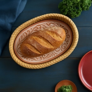 Fresh loaf of bread resting on a terracotta bread warmer in a woven basket, styled on a farmhouse table with herbs and dinnerware.