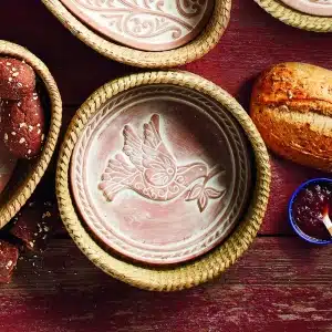 Terracotta bread warmer with carved dove design inside a woven basket, surrounded by fresh bread and jam on a rustic wooden table.